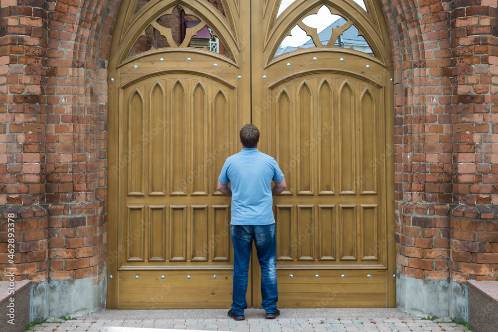 A tall man stands in front of the huge wooden doors of the church.
