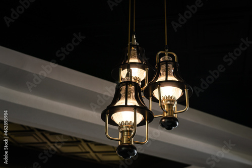A luxury celing lighting lamp is glowing in warm light shade, with beam structure and dark area as background. Interior decoration furniture object photo. Close-up and selective focus.