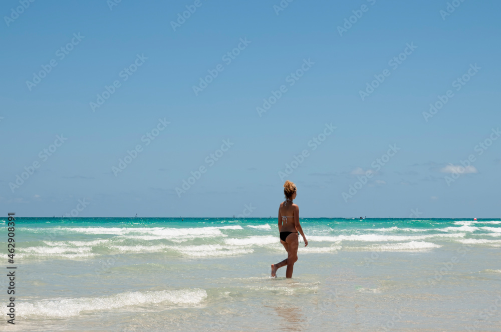 A woman with a fit body, wearing bikini walking on Miami beach on a summer day with a cloudless sky