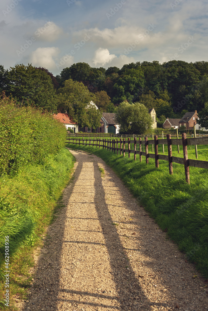 Unpaved footpath with a wooden fence along it in a sunny hilly landscape with houses and trees under a blue cloudy sky. Unpaved footpath with a wooden fence along it in a sunny hilly landscape with houses and trees under a blue cloudy sky.