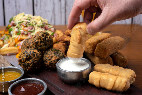 hand dipping a tequeño in white sauce on chopped board with tequeños, chicken sticks, vegetable croquettes with assorted sauces, beer and french fries