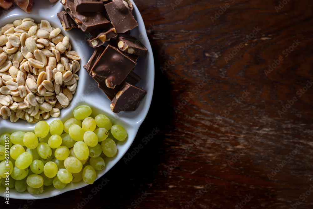 assorted appetizer on a plate shot from above. nuts, bacon, cheese, chocolate, grapes on a wooden background