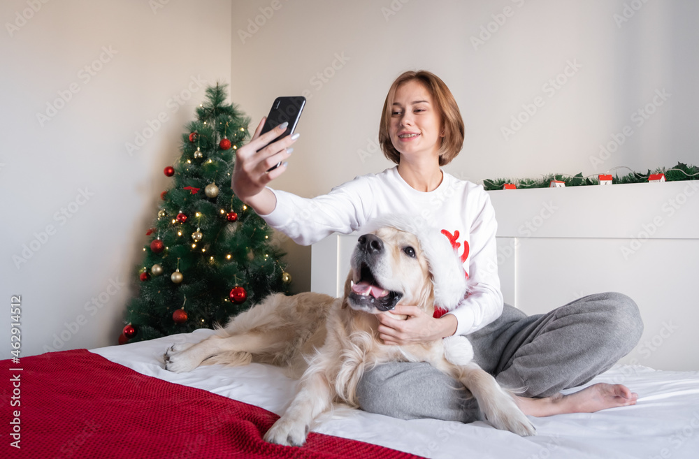 Portre of a young woman with a dog in a Christmas atmosphere. The girl looks at the phone in the hat of Santa Claus with a golden retriever on the bed near the Christmas tree. Portre of a young woman with a dog in a Christmas atmosphere. The girl looks at the phone in the hat of Santa Claus with a golden retriever on the bed near the Christmas tree.
