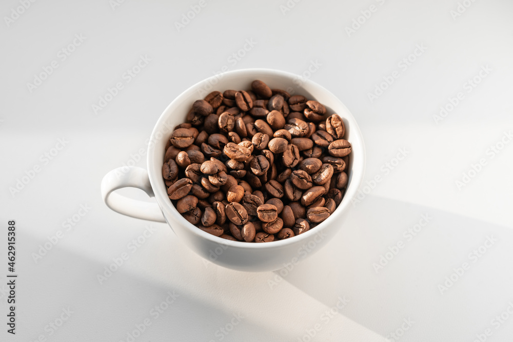 A white cup filled with brown coffee beans on white background A white cup filled with brown coffee beans on white background