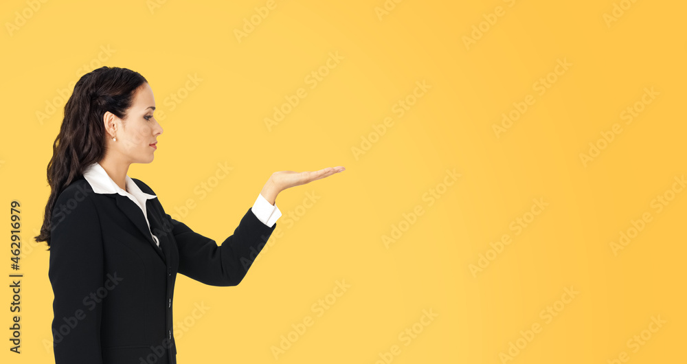 Profile side of beautiful businesswoman showing something or copy spase for product. Brunette woman looking at her hand in business studio concept, isolated over yellow background. Profile side of beautiful businesswoman showing something or copy spase for product. Brunette woman looking at her hand in business studio concept, isolated over yellow background.