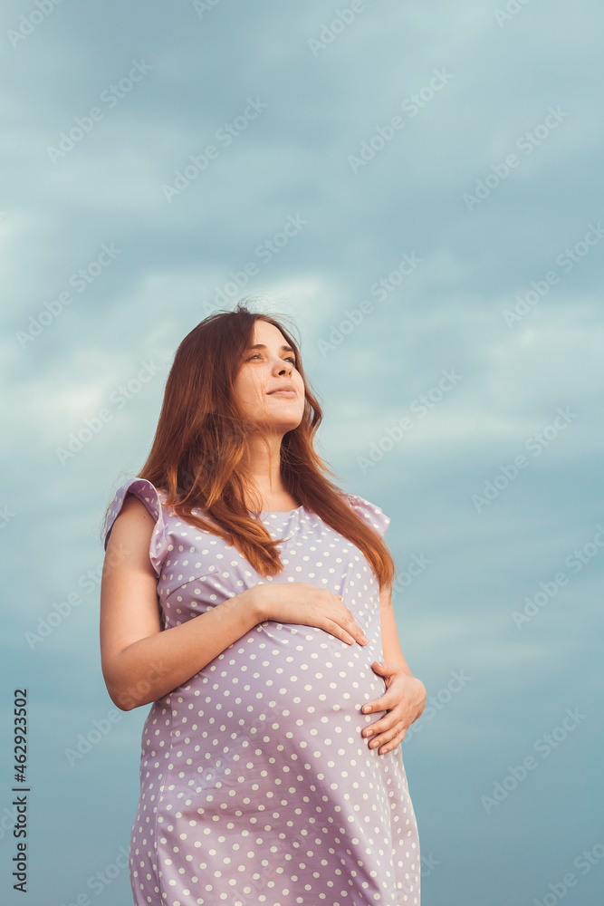 red-haired pregnant woman in dress stands against background of gloomy cloudy sky caress belly and looking up to the sky with gratitude