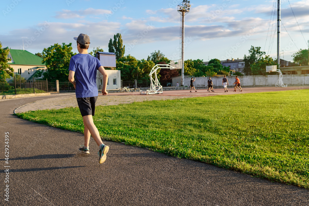 concept of sports and health - teen boy runs along the stadium track, a soccer field with green grass. - obrazy, fototapety, plakaty 
