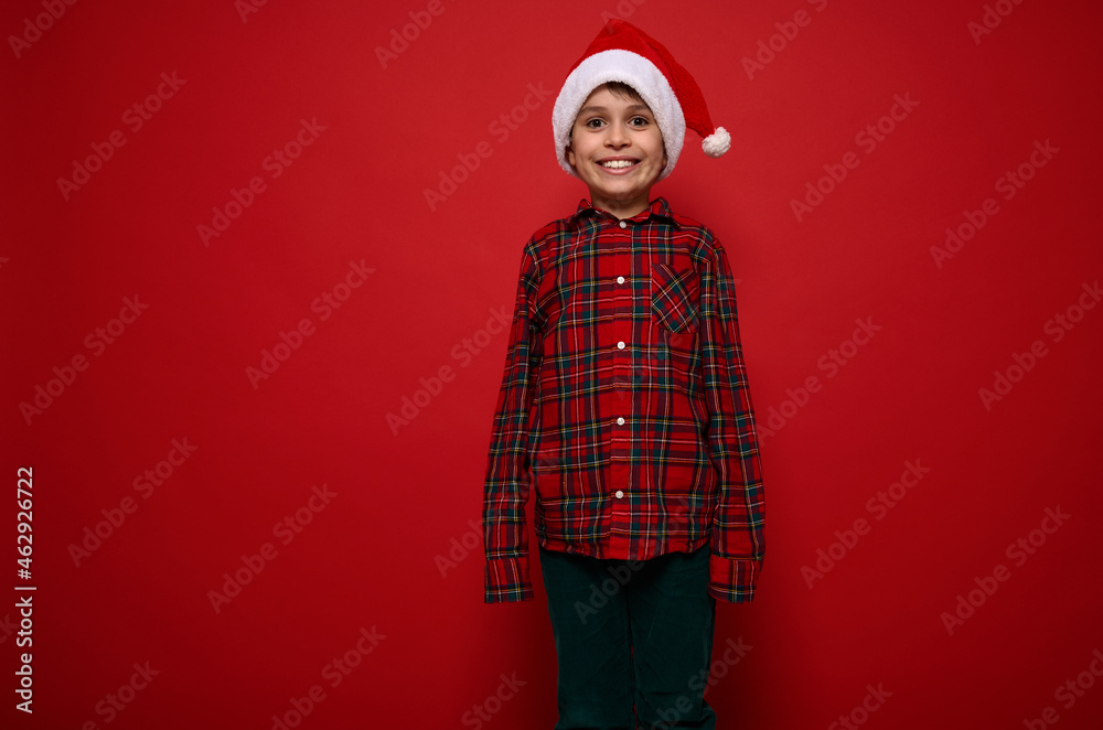Fototapeta premium Isolated portrait on colored background with copy space for Christmas and New Year ad of cheerful funny cute preadolescent boy in red plaid shirt and green velvet trousers smiling looking at camera