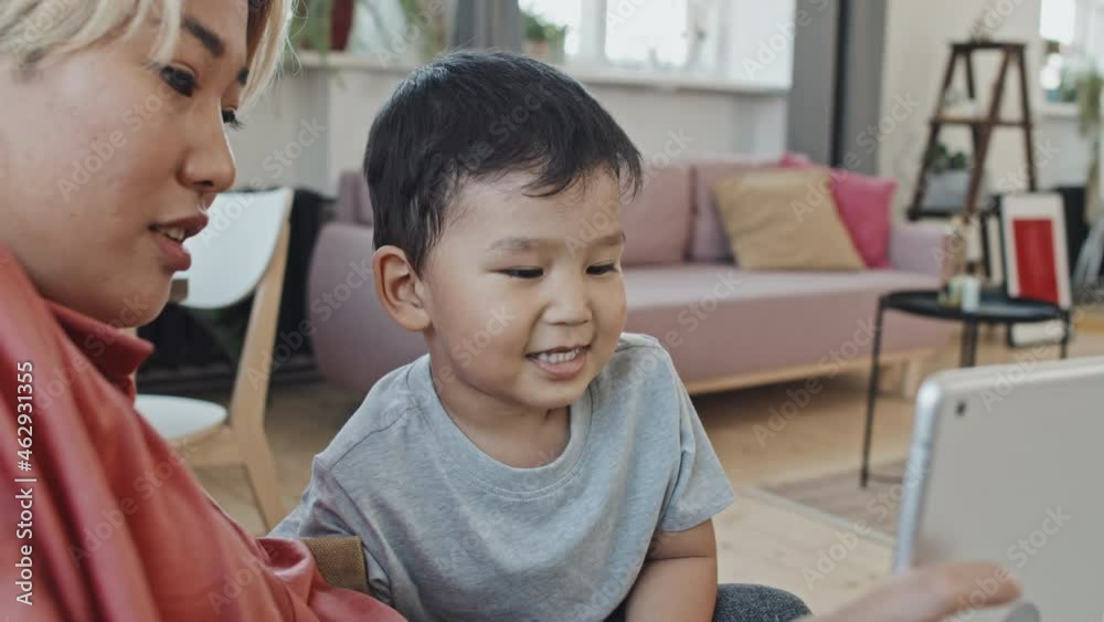 Medium close-up with slowmo of cute smiling 3-year-old Asian boy making funny faces looking at display of digital tablet in his mothers hands, spending weekend together at cozy home Medium close-up with slowmo of cute smiling 3-year-old Asian boy making funny faces looking at display of digital tablet in his mothers hands, spending weekend together at cozy home