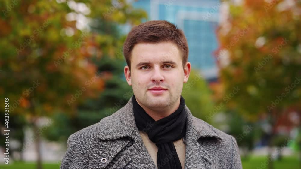 Young man wearing coat and scarf standing in autumn park and smiling at camera, building behind trees on blurred background. Portrait of city dweller outside. Concept of appearance Young man wearing coat and scarf standing in autumn park and smiling at camera, building behind trees on blurred background. Portrait of city dweller outside. Concept of appearance