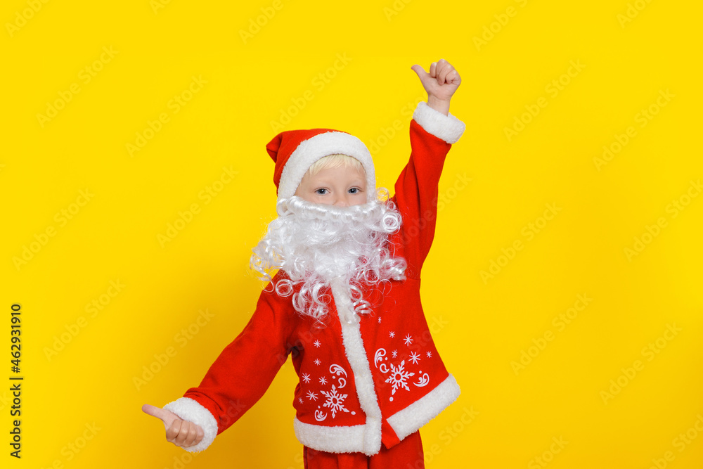 Three-year-old child in Santa Claus clothes and with a white beard shows a thumbs up gesture, stands on a yellow studio background. Three-year-old child in Santa Claus clothes and with a white beard shows a thumbs up gesture, stands on a yellow studio background.