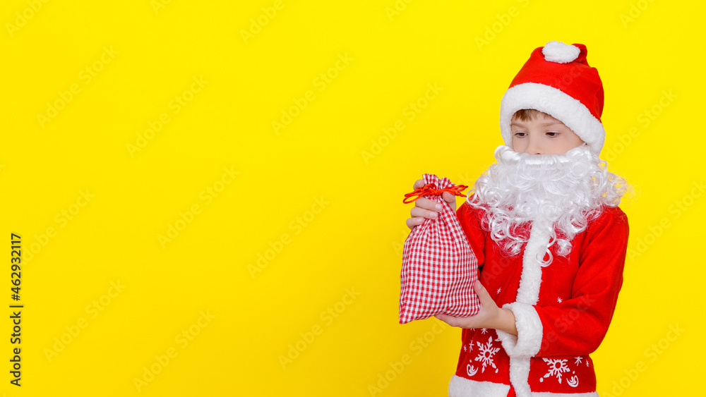 A child in Santa Claus clothes and an artificial beard holds a small bag with New Year's gifts in his hands, copy space, yellow studio background. A child in Santa Claus clothes and an artificial beard holds a small bag with New Year's gifts in his hands, copy space, yellow studio background.