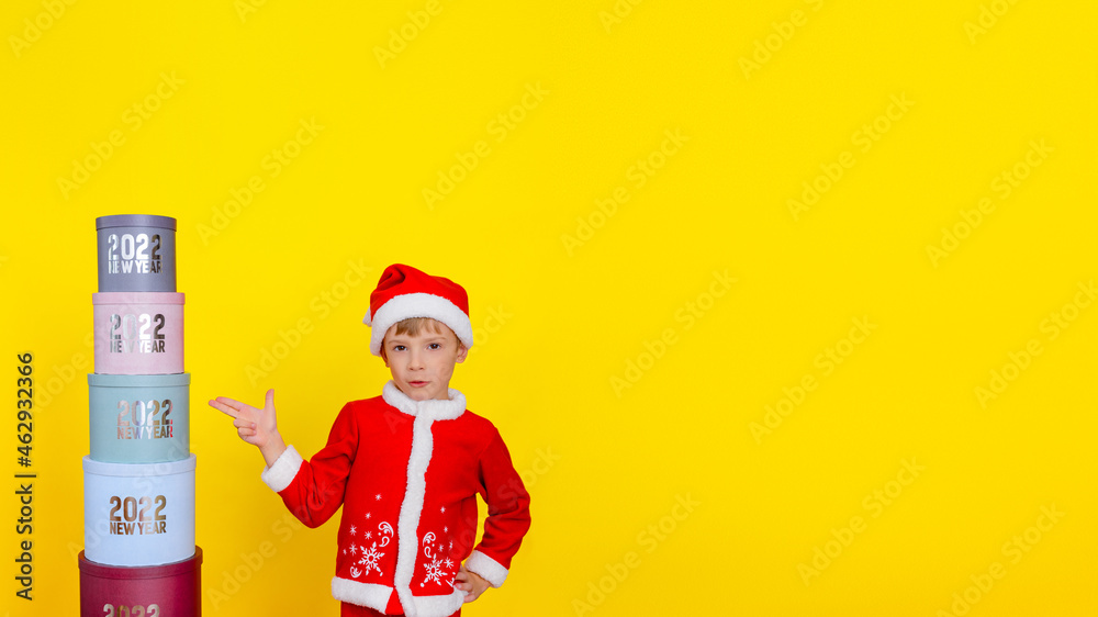 A six-year-old cute smiling child stands near a tower of round gift boxes with the inscriptions New Year 2022, shows a hand gun gesture, yellow background, copy space.
