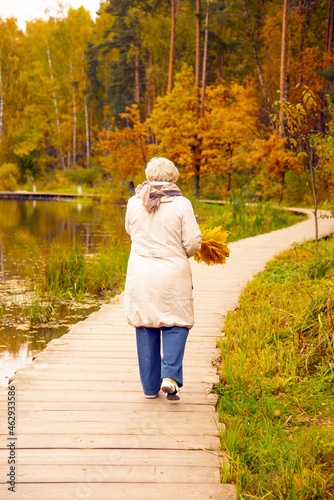 An elderly woman walks on a wooden deck over a pond in an autumn park.
