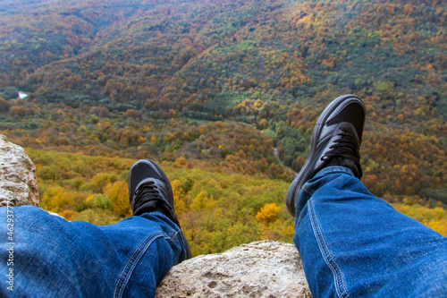 Male legs in sneakers sit on the edge of a high cliff and watch the mountains and forest on a sunny autumn day