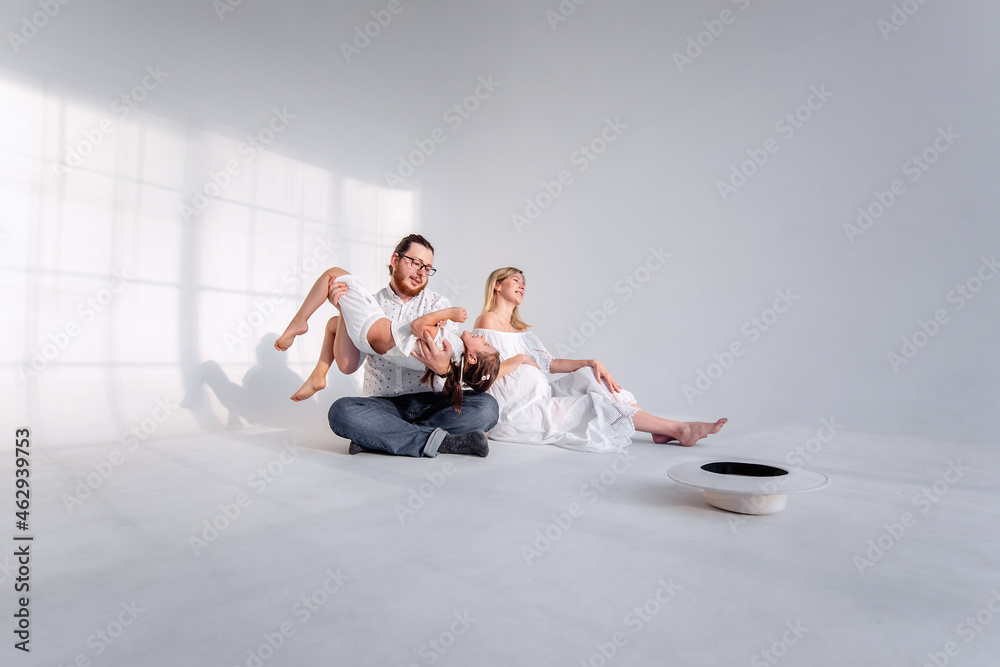 In white minimalistic interior, the family is having fun sitting on the floor. Young woman pregnant