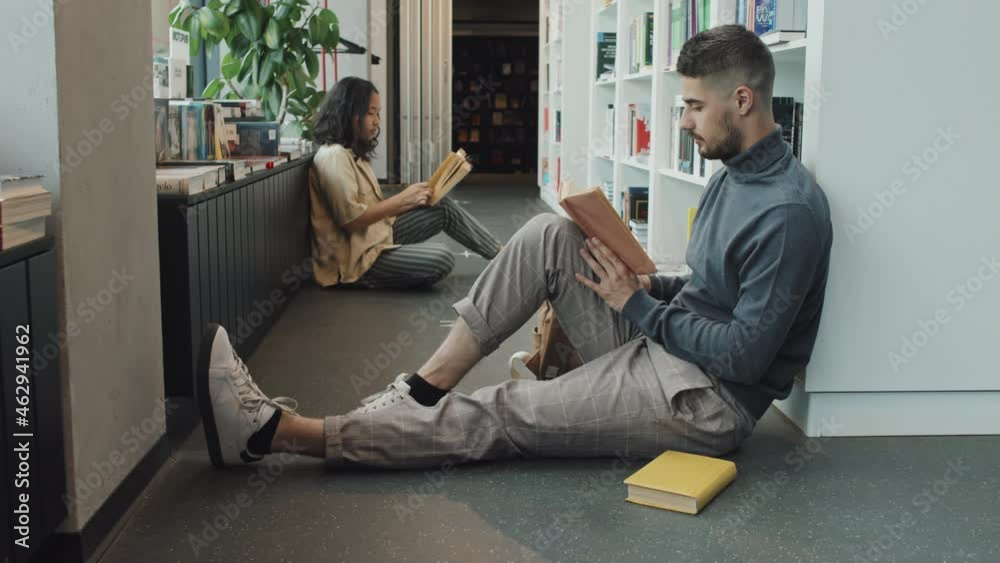 Stab slowmo shot of young multi-ethnic man and woman reading while sitting on floor next to bookcase in modern library Stab slowmo shot of young multi-ethnic man and woman reading while sitting on floor next to bookcase in modern library