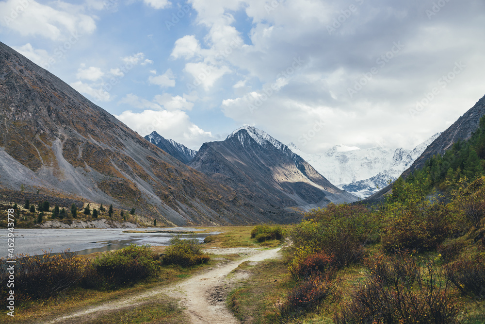 Scenic landscape with footpath along water streams in mountain valley in autumn colors with view to snowy mountains. Colorful autumn scenery with pathway near mountain creek and snow-covered mountains Scenic landscape with footpath along water streams in mountain valley in autumn colors with view to snowy mountains. Colorful autumn scenery with pathway near mountain creek and snow-covered mountains