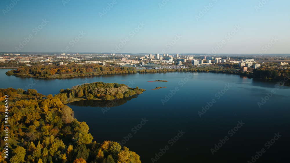 Flight over the autumn park. Park on the shore of a large lake. Trees with yellow autumn leaves are visible. On the horizon there is a blue sky and city houses. Aerial photography.