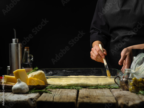 The chef greases the ready-to-bake glass dough with butter. Ingredients for making pie, pizza, focaccia. Black background, wood texture. Restaurant, hotel, bakery, pizzeria.