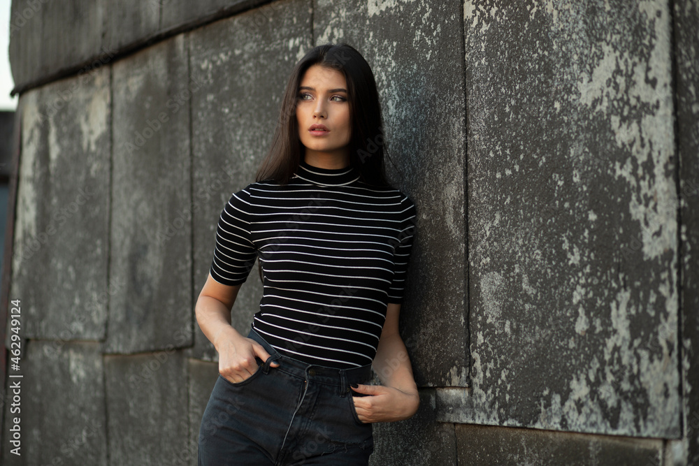Beautiful brunette girl in casual outfit with slim body, stand near old wall.
