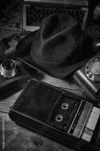 Old vintage cinematic noir scene, detective's desk with a hat, telephone, camera, portable cassette recorder, and whisky