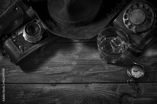 Old vintage cinematic noir scene, detective's desk with a hat, telephone, camera, portable cassette recorder, and whisky photo