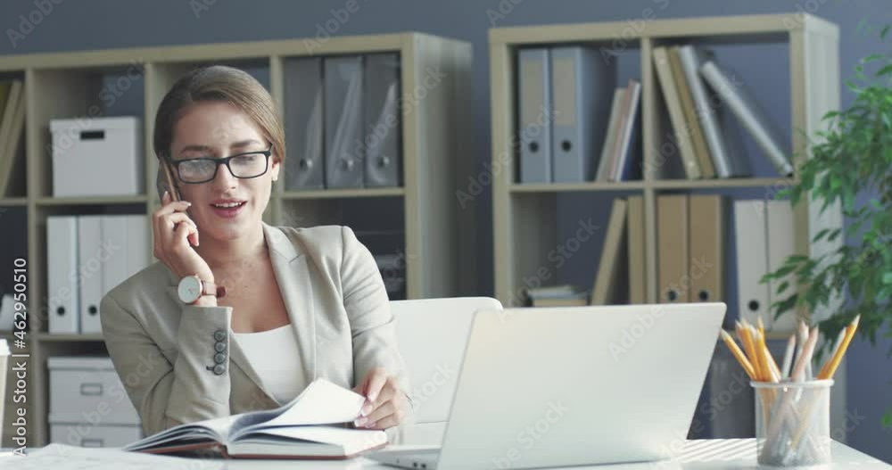 Young beautiful female office worker sitting at workplace and chatting on mobile phone looking through her paper business diary.