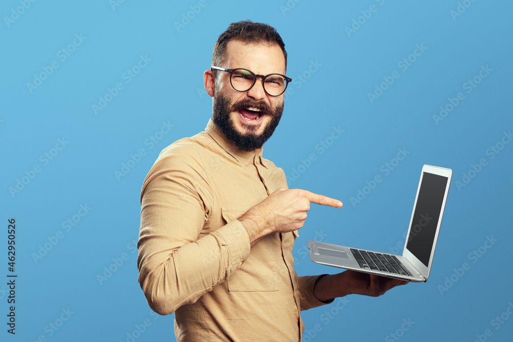 Trendy hipster man in beige shirt and eyeglasses screaming with excitement and pointing at new laptop Trendy hipster man in beige shirt and eyeglasses screaming with excitement and pointing at new laptop