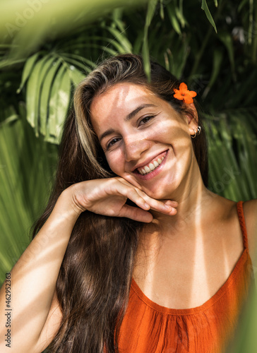 beautiful Hispanic woman in a terracotta dress with a flower in her ear smiles happily - hand on her face