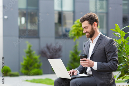 cheerful and successful male businessman working on laptop at lunchtime sitting on bench near office in business suit and drinking hot drink, banker near office