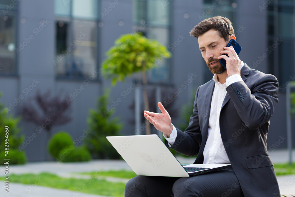 Successful male businessman reports good news by phone, working with laptop at lunchtime near office sitting on bench Successful male businessman reports good news by phone, working with laptop at lunchtime near office sitting on bench
