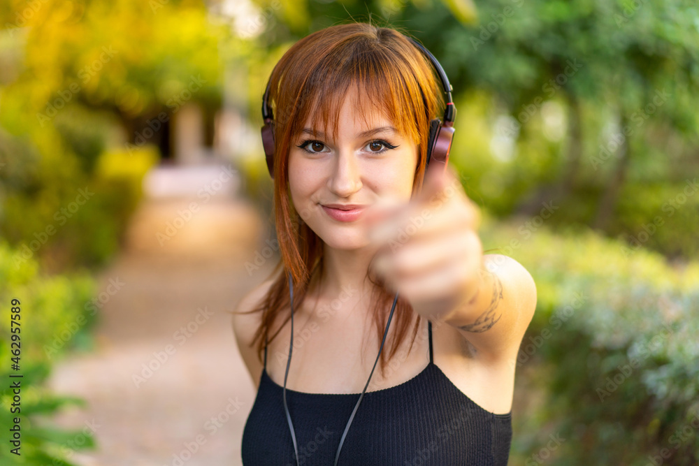 Young pretty redhead woman at outdoors listening music and pointing to the front