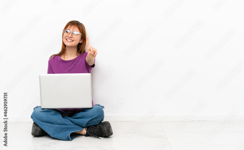 redhead girl with a laptop sitting on the floor showing and lifting a finger redhead girl with a laptop sitting on the floor showing and lifting a finger