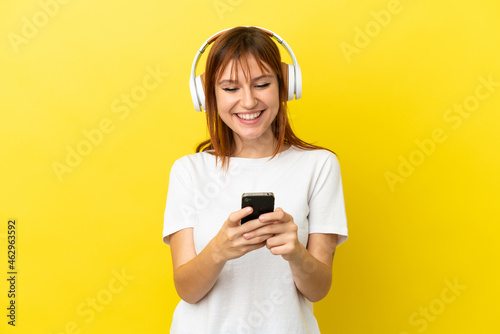 Redhead girl isolated on yellow background listening music and looking to mobile