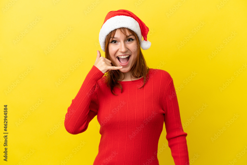 Redhead girl with christmas hat isolated on yellow background making phone gesture. Call me back sign Redhead girl with christmas hat isolated on yellow background making phone gesture. Call me back sign