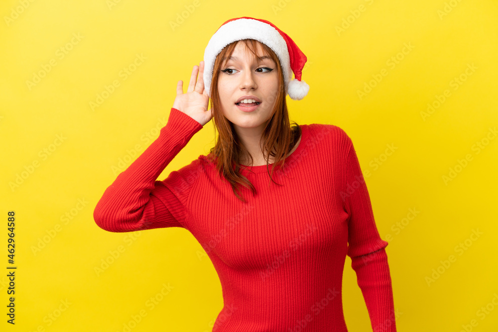 Redhead girl with christmas hat isolated on yellow background listening to something by putting hand on the ear Redhead girl with christmas hat isolated on yellow background listening to something by putting hand on the ear