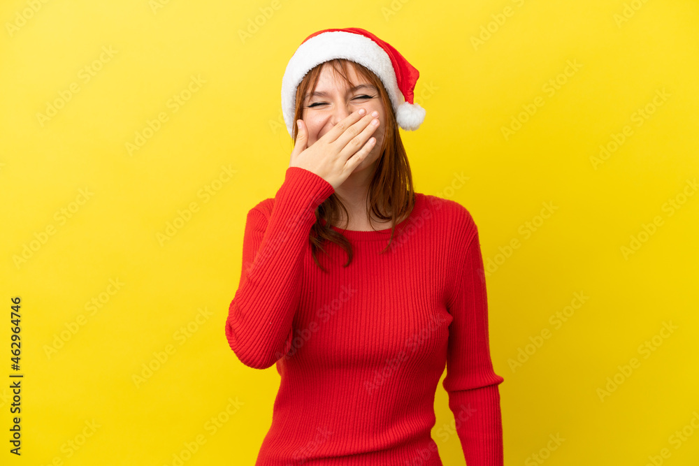 Redhead girl with christmas hat isolated on yellow background happy and smiling covering mouth with hand