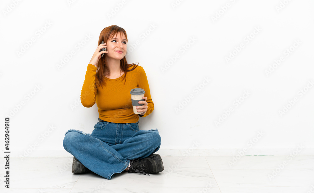Redhead girl sitting on the floor isolated on white background holding coffee to take away and a mobile