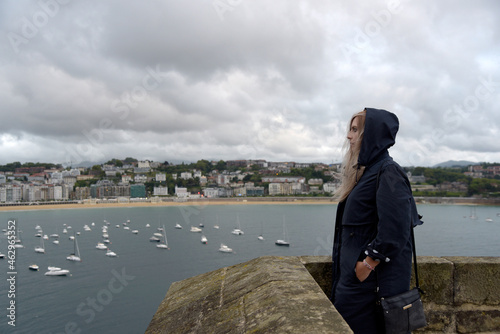 Una mujer rubia con ropa oscura de costado mirando hacia el mar en San Sebastian con el cielo lleno de nubes