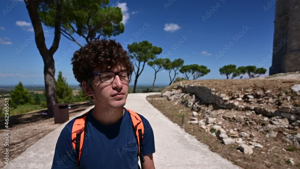 Andria, Puglia, Italy. August 2021. Footage with the portrait of a Caucasian young man with glasses walking along the road around Castel del Monte observing it. Final shot on the castle.