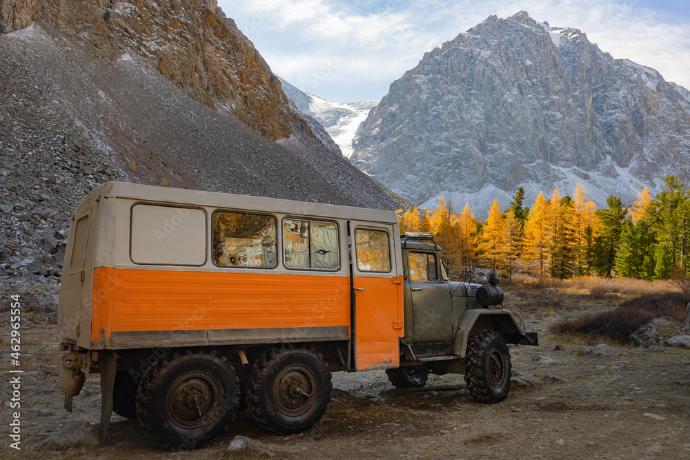 The off-road truck stands against the background of mountains, yellow larches and a glacier. Tourism and travel. Russia, Altai.