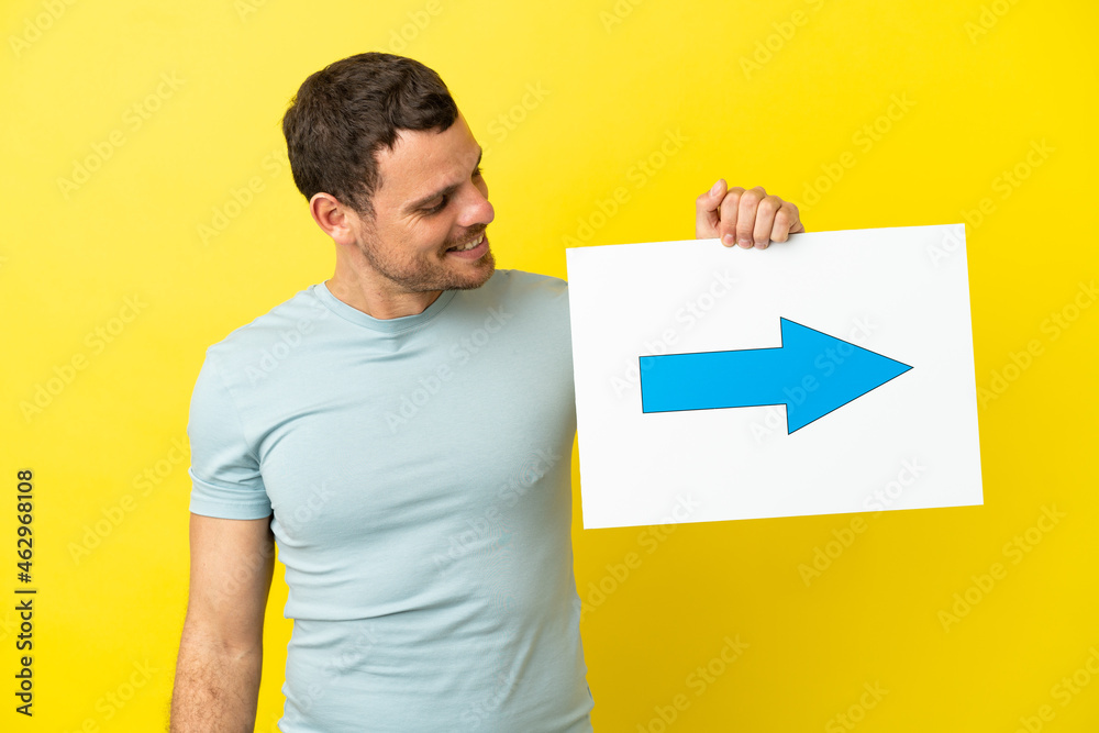 Brazilian man over isolated purple background holding a placard with arrow symbol Brazilian man over isolated purple background holding a placard with arrow symbol