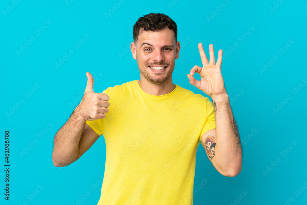 Fototapeta premium Young caucasian handsome man isolated on blue background showing ok sign and thumb up gesture