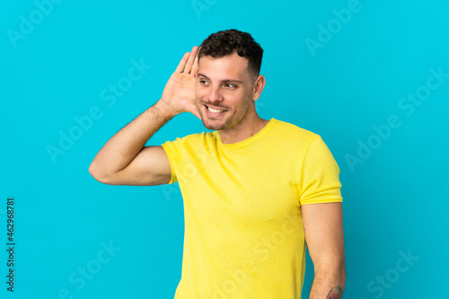 Young caucasian handsome man isolated on blue background listening to something by putting hand on the ear