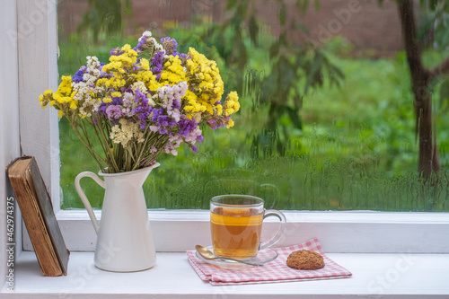 Old book, colorful bouquet of flowers and white cup of tea on background of window with raindrops at home at summer day