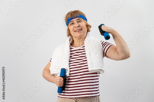 An elderly woman with a blue bandage on her head trains with dumbbells on a white background © Михаил Решетников