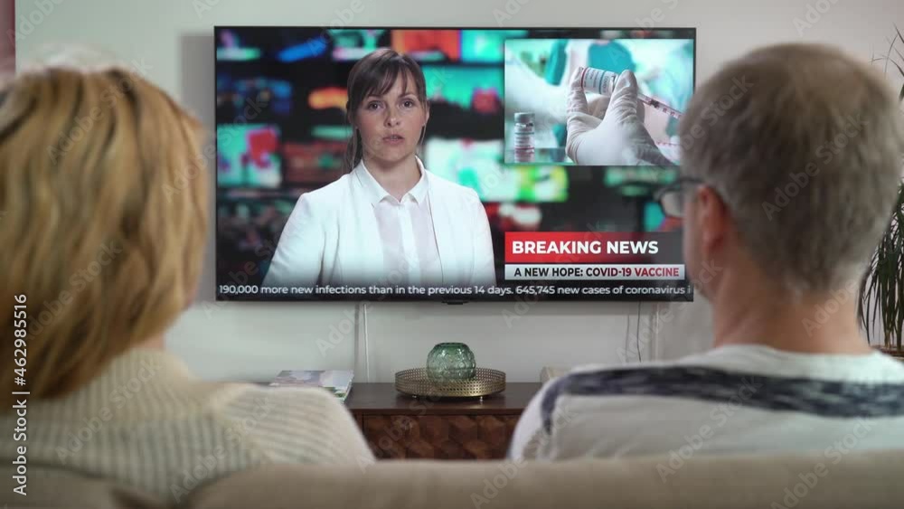 Family Couple Watching TV News Sitting on Couch in Living Room Together. TV Presenter Telling Breaking News about Coronavirus, Lockdown and Vaccinations. Rear View Family Couple Watching TV News Sitting on Couch in Living Room Together. TV Presenter Telling Breaking News about Coronavirus, Lockdown and Vaccinations. Rear View