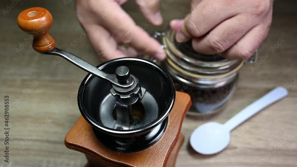 Hands opening a jar, uses a white spoon to bring beans into the grinder and returns the spoon on the table; Coffee Grinder, Jar with Coffee Beans, Spoon; Hands opening a jar, uses a white spoon to bring beans into the grinder and returns the spoon on the table; Coffee Grinder, Jar with Coffee Beans, Spoon;