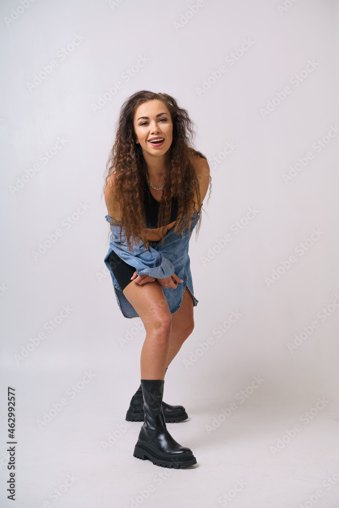 Cheerful young woman is dancing on a light background in a denim jacket and black sports shorts and boots. Caucasian brunette girl posing on background in studio Cheerful young woman is dancing on a light background in a denim jacket and black sports shorts and boots. Caucasian brunette girl posing on background in studio